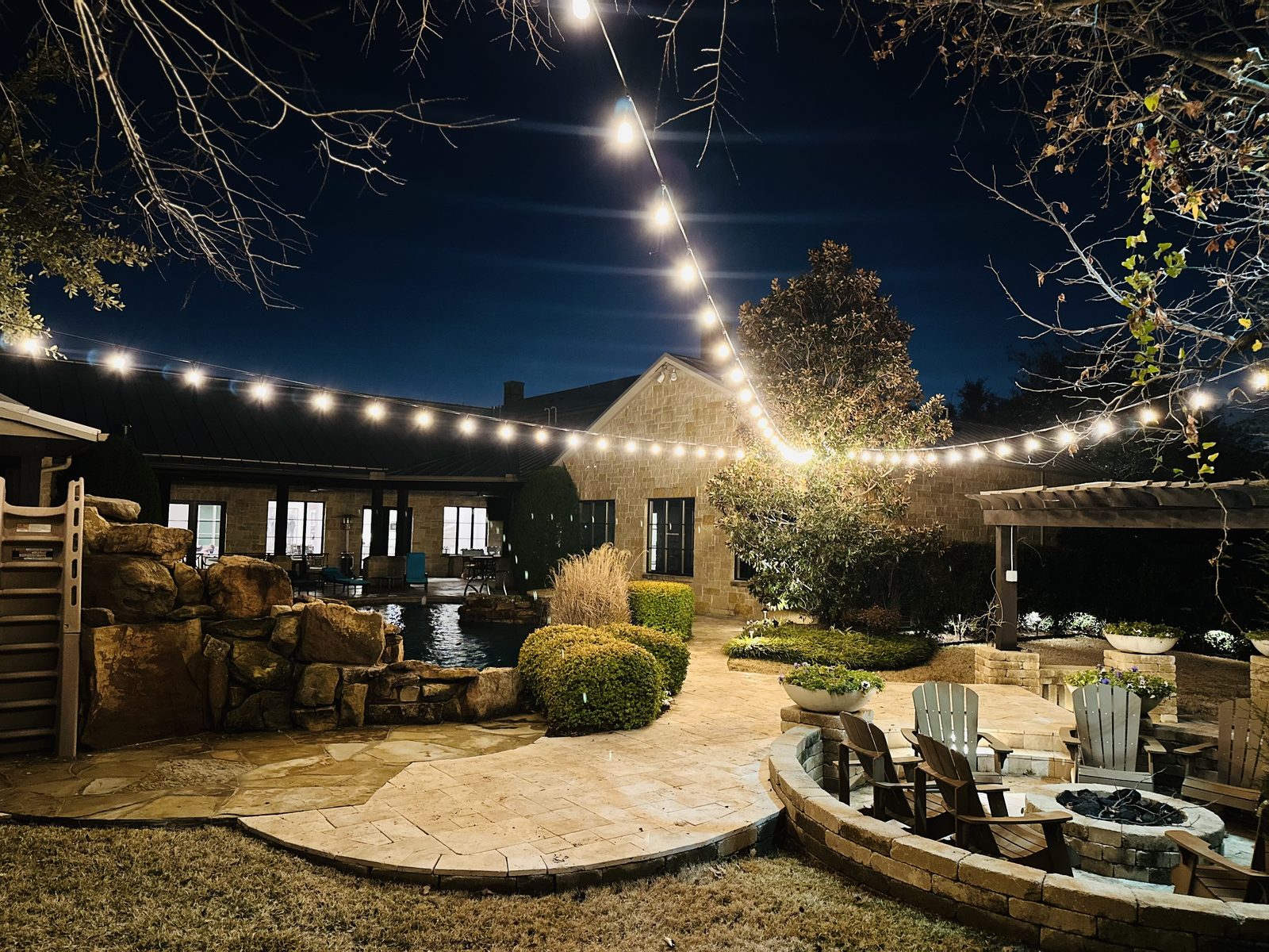 Backyard patio at night with overhead string lighting, a fire pit and a lit stone water feature