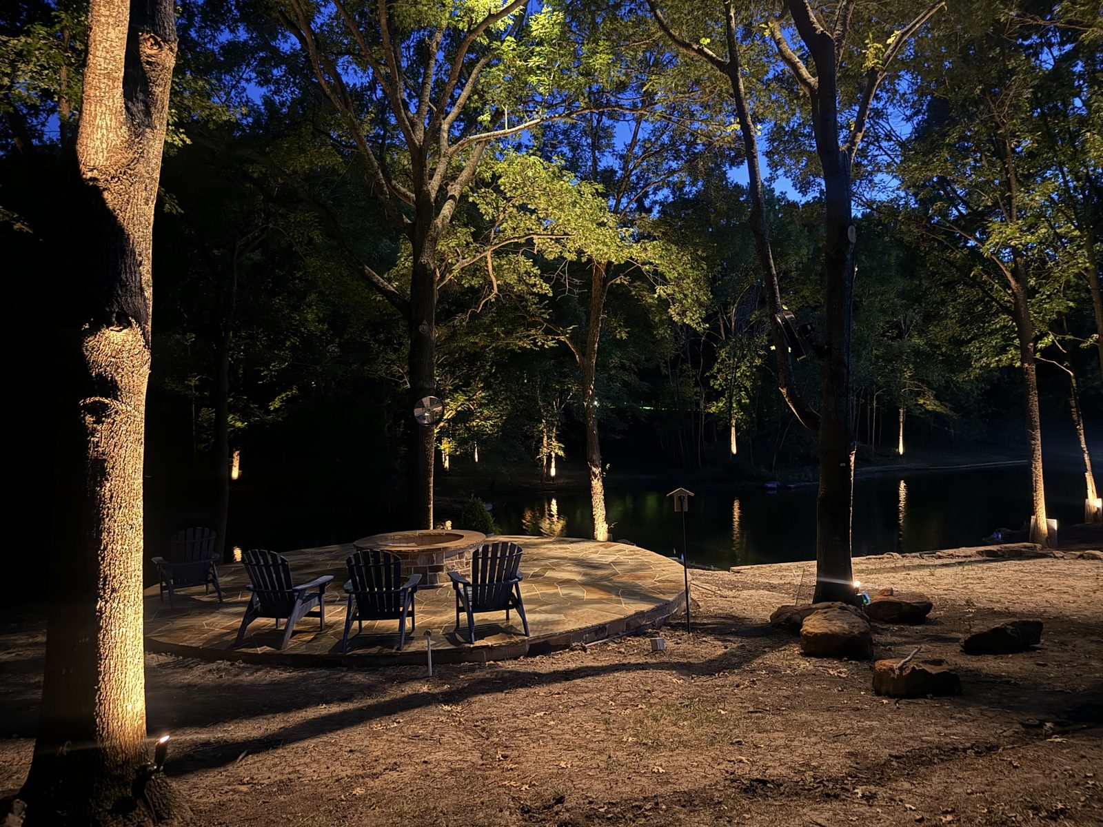 Lakeside seating area at night under uplit trees with a table and chairs on the shoreline